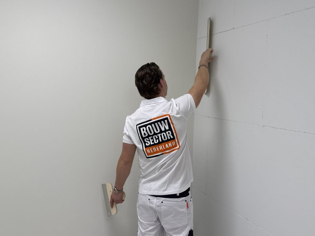 Stukadoor werkt met een lange spaan de hoek van een wand bij, met een kleine spaan in zijn andere hand
